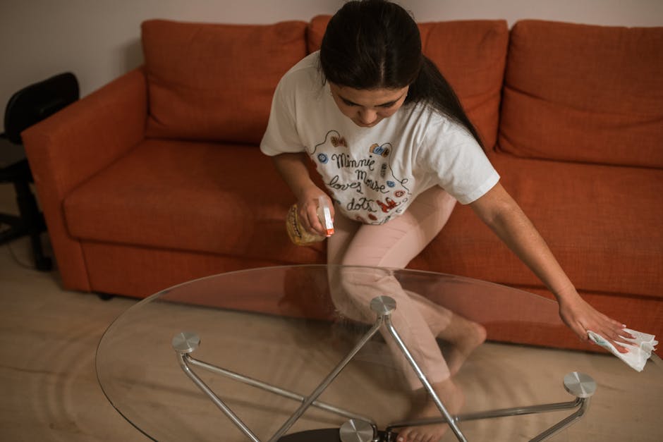 A young woman wearing a lavender sweatshirt and yellow cleaning gloves is kneeling on a dark wooden floor, polishing a round, glass-topped side table with a pink cleaning cloth. The room features mode