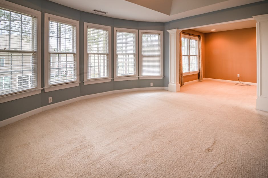 A spacious residential living room with light beige carpet flooring and a soft pink accent wall. The room features a large set of white-framed windows with white blinds, allowing natural light to illu