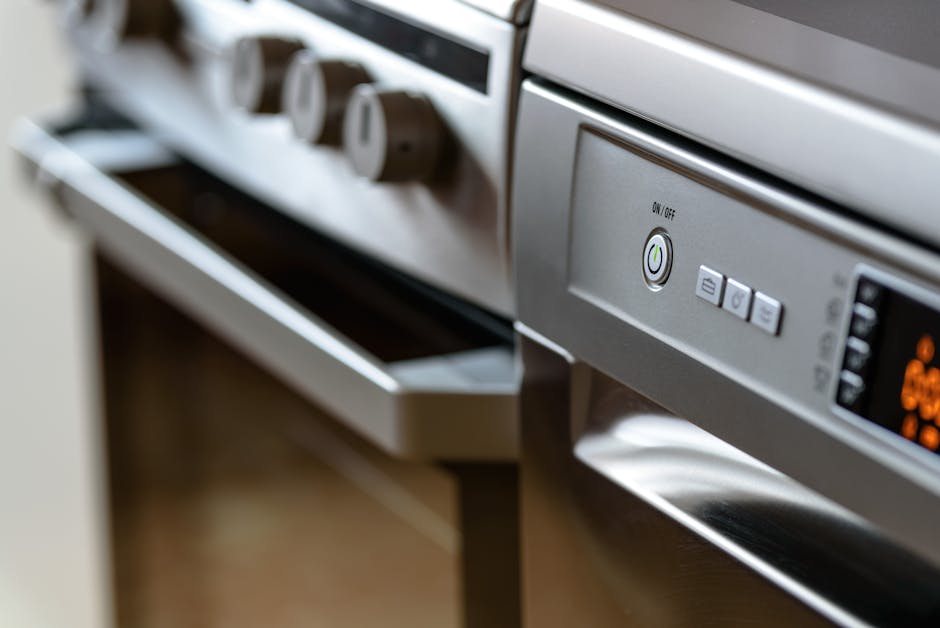 A modern kitchen featuring a white under-counter oven with a stainless steel handle and control panel, surrounded by white cabinetry with sleek, flat-front doors. The grey countertop extends across th
